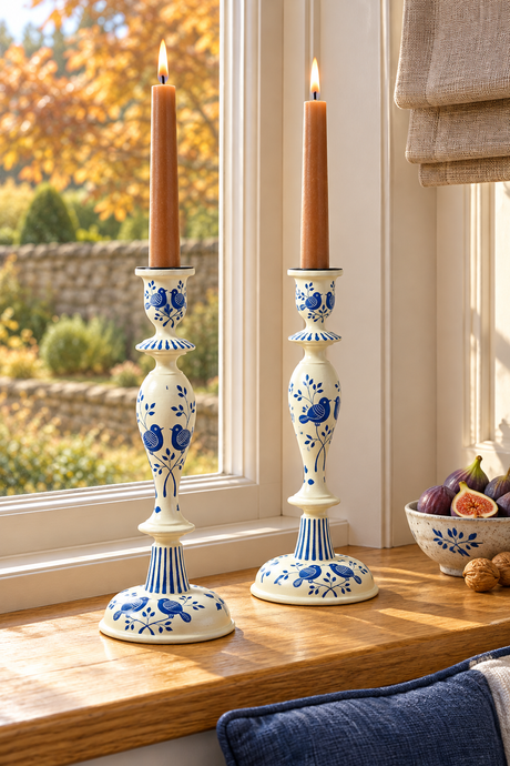 Two tall wood candlesticks with blue floral patterns and lit taper candles, displayed on a wooden windowsill in a bright room.