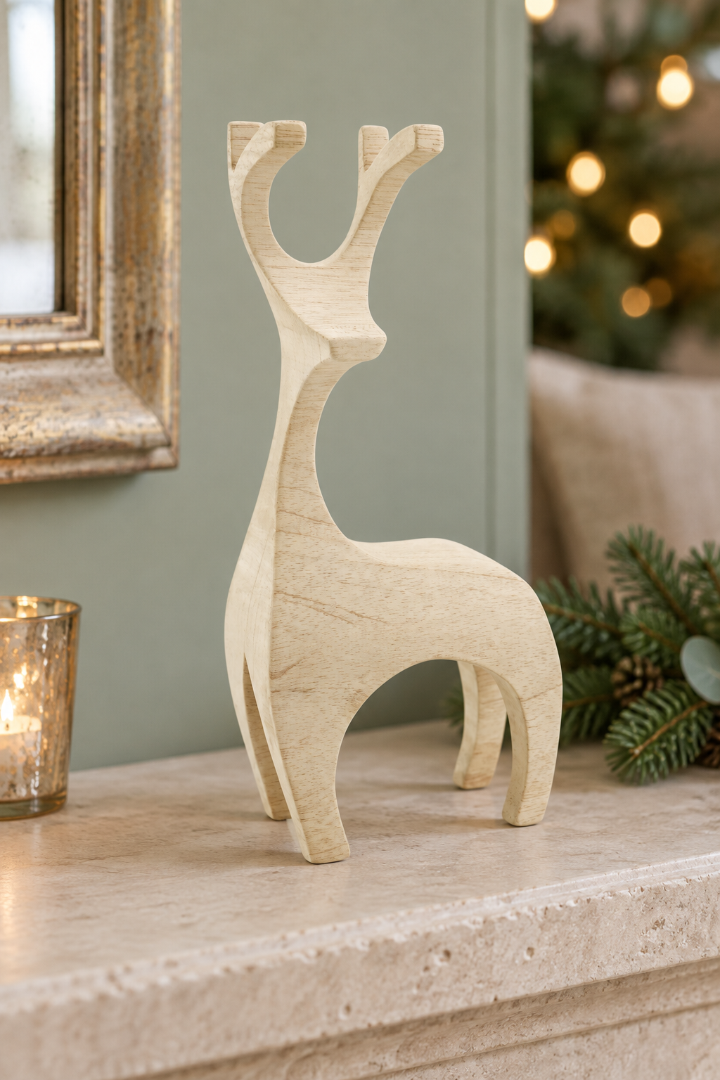 A light natural wood reindeer figurine standing on a stone mantelpiece, with a lit candle holder, mirror frame, pine branches, and a softly blurred Christmas tree in the background.