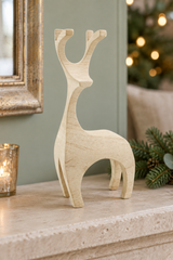 A light natural wood reindeer figurine standing on a stone mantelpiece, with a lit candle holder, mirror frame, pine branches, and a softly blurred Christmas tree in the background.