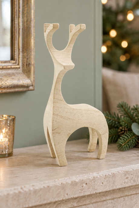 A light natural wood reindeer figurine standing on a stone mantelpiece, with a lit candle holder, mirror frame, pine branches, and a softly blurred Christmas tree in the background.