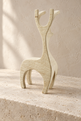 A minimalist, sculptural wooden deer figurine in a light natural wood finish, standing on a textured stone ledge against a warm beige plaster wall.