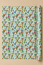A roll of wrapping paper with a colorful repeating Patchwork Giraffe pattern on a light blue background, displayed against a solid beige backdrop.