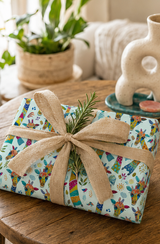A wrapped gift with a colourful patchwork giraffe-patterned paper and a large tan burlap ribbon bow, tied with a small green sprig, resting on a rustic wooden table in a cozy indoor setting.