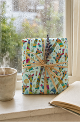 A gift box wrapped in a colourful giraffe-patterned paper with a natural twine bow and a sprig of lavender, sitting on a windowsill next to a steaming mug and an open book.