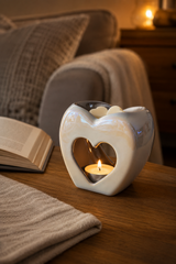A heart-shaped Pearl Heart Wax Melt Burner ceramic candle holder with a lit tea light candle inside, placed on a wooden table in a cozy living room setting with a sofa and book in the background.
