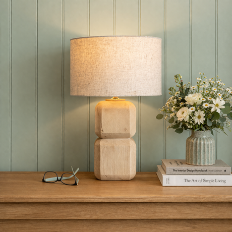 Wooden lamp on a wooden surface with books and flowers against a green paneled wall.