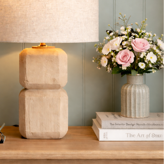 Beige table lamp on a wooden surface with books and flowers in the background.