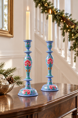 Two tall blue ceramic candlestick holders with floral hand-painted details, each holding a lit white taper candle, displayed on a wooden console table in a decorated indoor staircase setting.