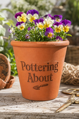 An orange-brown terracotta planter pot with the words 'Pottering About' and a small trowel graphic printed on the front, filled with purple, yellow, and white pansies. The pot sits on a rustic wooden surface with gardening props like scissors and a broken pot in a garden setting.