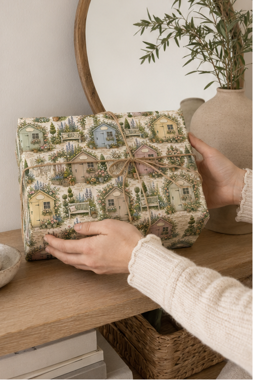 A person holds a square gift box wrapped in a Personalised Garden Shed Wrapping Paper with a small twine bow, set on a wooden shelf next to a vase with greenery.