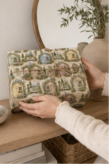 A person holds a square gift box wrapped in a Personalised Garden Shed Wrapping Paper with a small twine bow, set on a wooden shelf next to a vase with greenery.