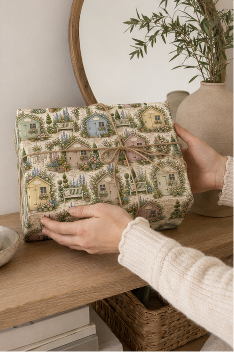 A person holds a square gift box wrapped in a Personalised Garden Shed Wrapping Paper with a small twine bow, set on a wooden shelf next to a vase with greenery.