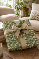 A square gift box wrapped in Personalised Hedgehog Garden Wrapping Paper with a natural burlap ribbon and a sprig of greenery, set on a wooden table in a cozy living room.