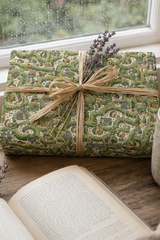 A rectangular gift box wrapped in Personalised Hedgehog Garden Wrapping Paper  with a natural twine bow and lavender sprig, placed on a wooden surface near an open book and a window.