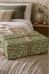 A rectangular gift box wrapped in Personalised Hedgehog Garden Wrapping Paper, tied with a natural twine bow, placed on a beige textured blanket in a softly lit bedroom setting.