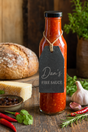 A glass bottle of Dan's Fire Sauce filled with red sauce, featuring a black tag with white text, styled on a rustic wooden table with bread, cheese, garlic, and red chilies.