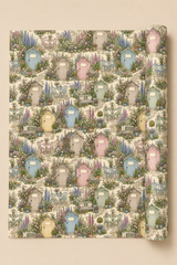 A rolled sheet of wrapping paper or gift wrap featuring a repeating pattern of pastel beach huts with door signs that can be personalised, benches, and garden foliage on a light cream background, shown against a solid beige backdrop.
