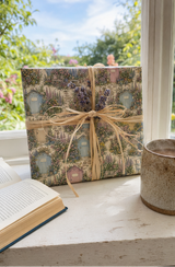 A gift box wrapped in a vintage-style Personalised Pastel Shed Village Wrapping Paper, tied with a natural jute ribbon, sitting on a white windowsill next to an open book and a ceramic mug.