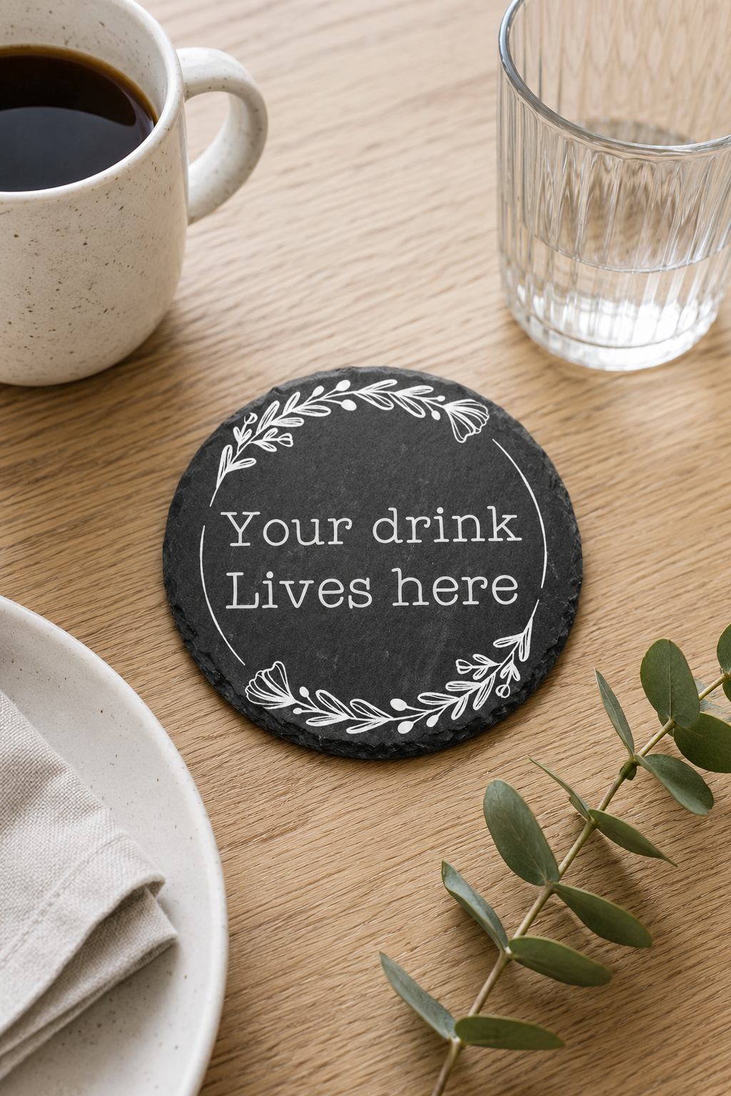 Coaster with 'Your drink lives here' text on a wooden table with a cup of coffee and glass.