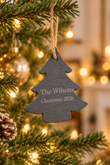 A dark gray slate Christmas tree ornament hanging from a pine tree branch with warm bokeh lights in the background. The ornament is personalized with the text 'The Wilsons Christmas 2026' in white lettering.