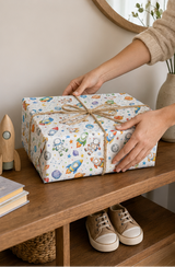 A gift box wrapped in white paper with a colorful space-themed print of astronauts, rockets, and planets, tied with a natural jute twine bow. The box is placed on a wooden shelf in a cozy room setting with a person's hands adjusting the twine.