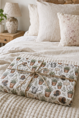 Personalised Terracotta Pot Wrapping Paper A wrapped rectangular gift box tied with twine sits on a bed. The wrapping paper has a botanical and gardening print with potted plants, watering cans, and small greenery illustrations on a light background.