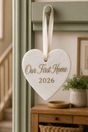A personalised marble heart-shaped white stone ornament hanging from a green door frame, featuring the engraved text 'Our First Home 2026' in brown lettering, with a cream ribbon loop at the top.