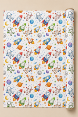 A roll of wrapping paper  with a colourful space-themed pattern for personalisation featuring cartoon rockets, astronauts, planets, moons, and stars on a white background, shown against a beige backdrop.