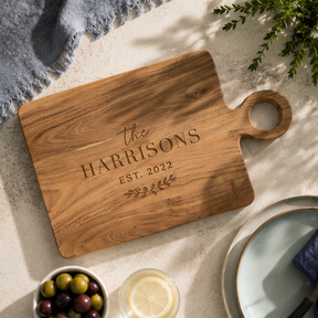 Wooden cutting board with 'the Harrisons Est. 2022' engraving on a stone surface.