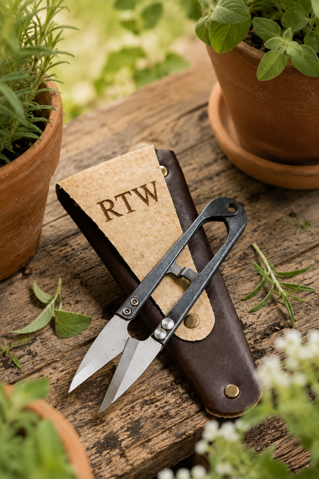 A personalised pair of small metal garden shears with a dark handle rests on a brown leather sheath marked RTW, placed on a rustic wooden surface with potted plants around it.
