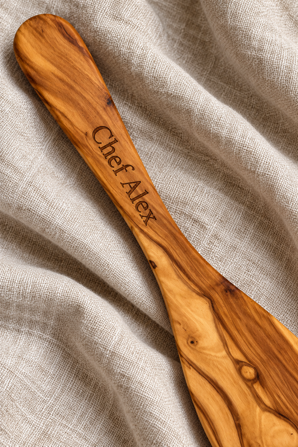 Close-up of “Chef Alex” laser engraved along an olive wood spatula handle, resting on natural linen.