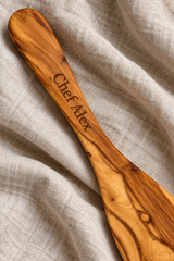 Close-up of “Chef Alex” laser engraved along an olive wood spatula handle, resting on natural linen.