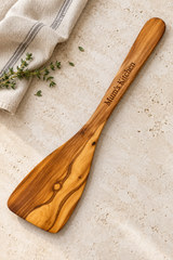 Solid olive wood spatula engraved “Mum’s Kitchen”, resting on pale stone beside striped linen and thyme.