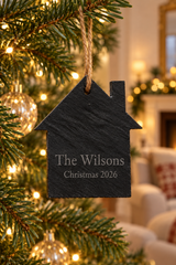 A personalized black slate Christmas ornament shaped like a house, hanging from a green Christmas tree with warm lights. The ornament is engraved with the name 'The Wilsons' and 'Christmas 2026'.