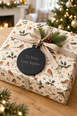 A Christmas-themed gift box wrapped in cream paper with a robin and holly pattern, tied with a beige ribbon and a pine sprig. A round black tag reads 'To Mum Love Sophie'.