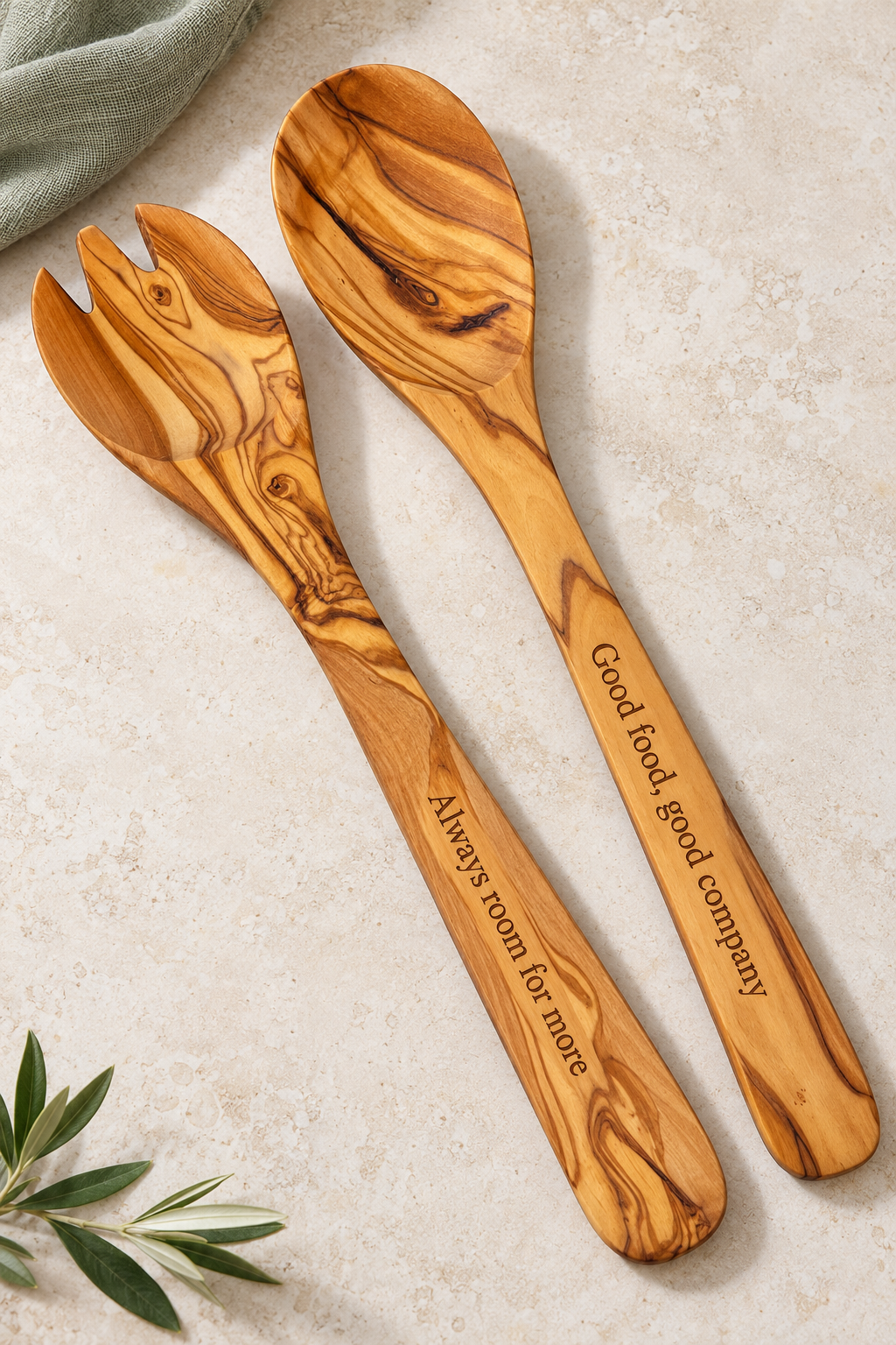 Medium olive wood salad spoon and fork engraved with “Good food, good company” and “Always room for more”, arranged on pale stone beside sage linen.
