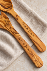 Close-up of “Alex & Jamie” laser engraved along both olive wood salad-server handles, showing the lettering and contrasting natural grain.