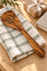 Olive wood spatula engraved “Made with Love”, styled on a checked tea towel beside a wrapped gift and recipe book.