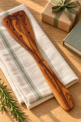 Olive wood spatula engraved “Made with Love”, styled on striped linen beside a wrapped gift and recipe book.