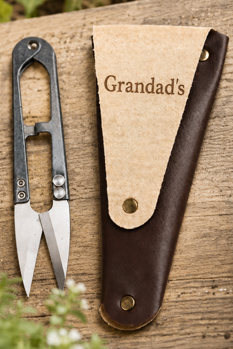 A personalised pair of metal garden snips with a dark handle and silver blades, placed beside a brown leather sheath on a rustic wooden surface. The sheath is printed with the name 'Grandad's' in brown lettering.