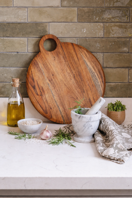 Wooden cutting board on a kitchen counter with herbs, oil, and a mortar and pestle.