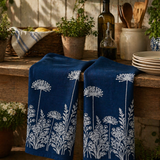 Blue hand printed linen tea towels with white floral pattern on a wooden table with plants and kitchen items.