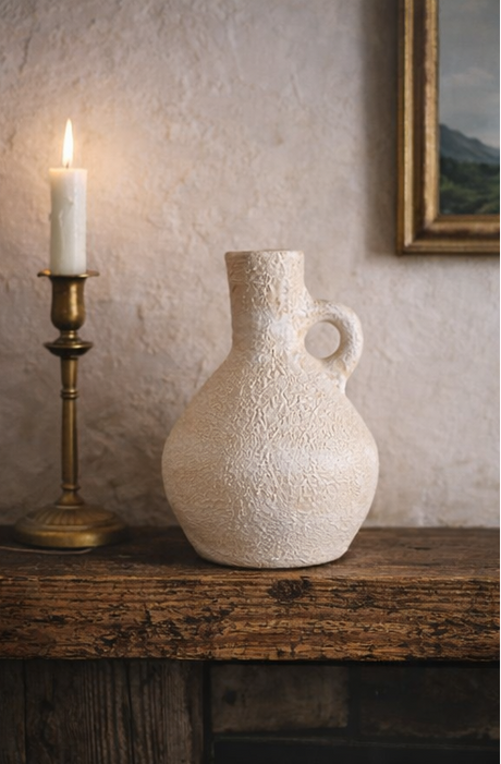 Textured ceramic vase on a wooden surface with a candle and framed picture in the background.