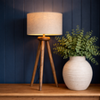 Wooden tripod lamp with beige shade and white vase with green plant against a dark blue wall.