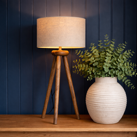 Wooden tripod lamp with beige shade and white vase with green plant against a dark blue wall.