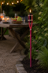 A lit pink bamboo-style garden torch light stands in a garden bed beside a wooden dining table at night, with blurred greenery and warm string lights in the background.