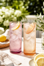 Two tall glasses with pink and orange hues filled with a sparkling drink, garnished with lemon slices, on a table outdoors.