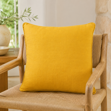 Plain mustard linen cushion cover with piped edge styled on a wicker chair.