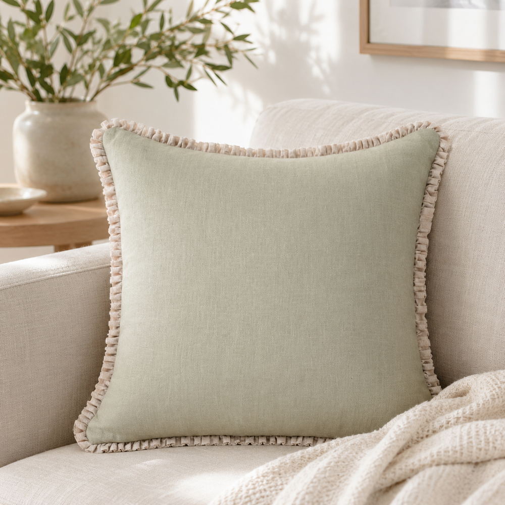 Plain sage linen cushion cover with pale ruffle edge styled on a neutral sofa by a window.