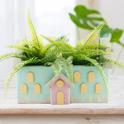 A decorative pastel-colored house plant holder with a pink felt base, filled with artificial green plants, placed on a wooden surface.